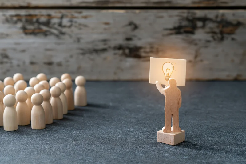 Wooden figurine holding glowing lightbulb sign facing a group of identical figures on textured surface