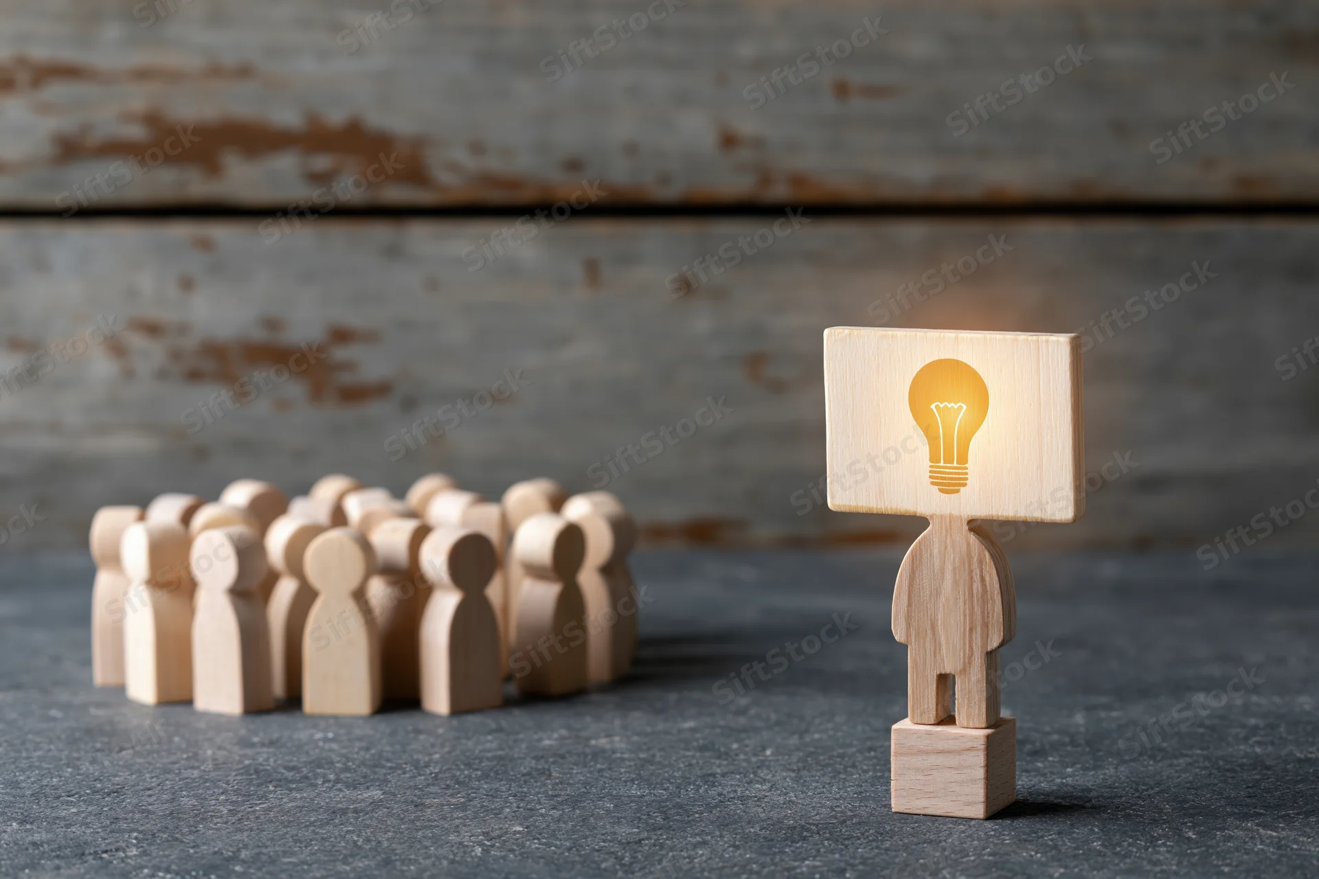 Wooden figurine with glowing lightbulb head standing apart from a group on a gray surface