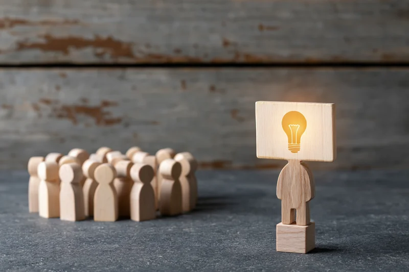 Wooden figurine with glowing lightbulb head standing apart from a group on a gray surface