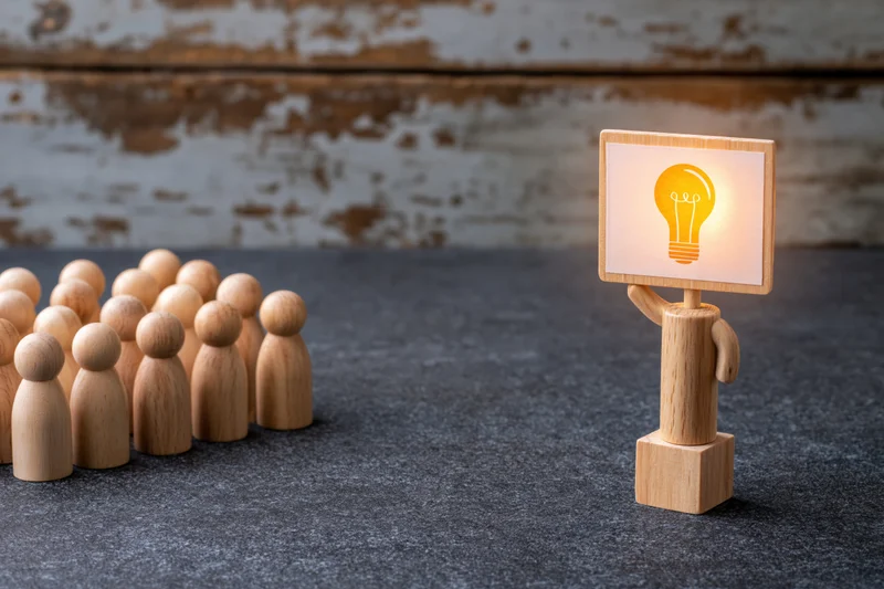 Wooden figurine holding illuminated lightbulb sign standing apart from a group on a gray surface