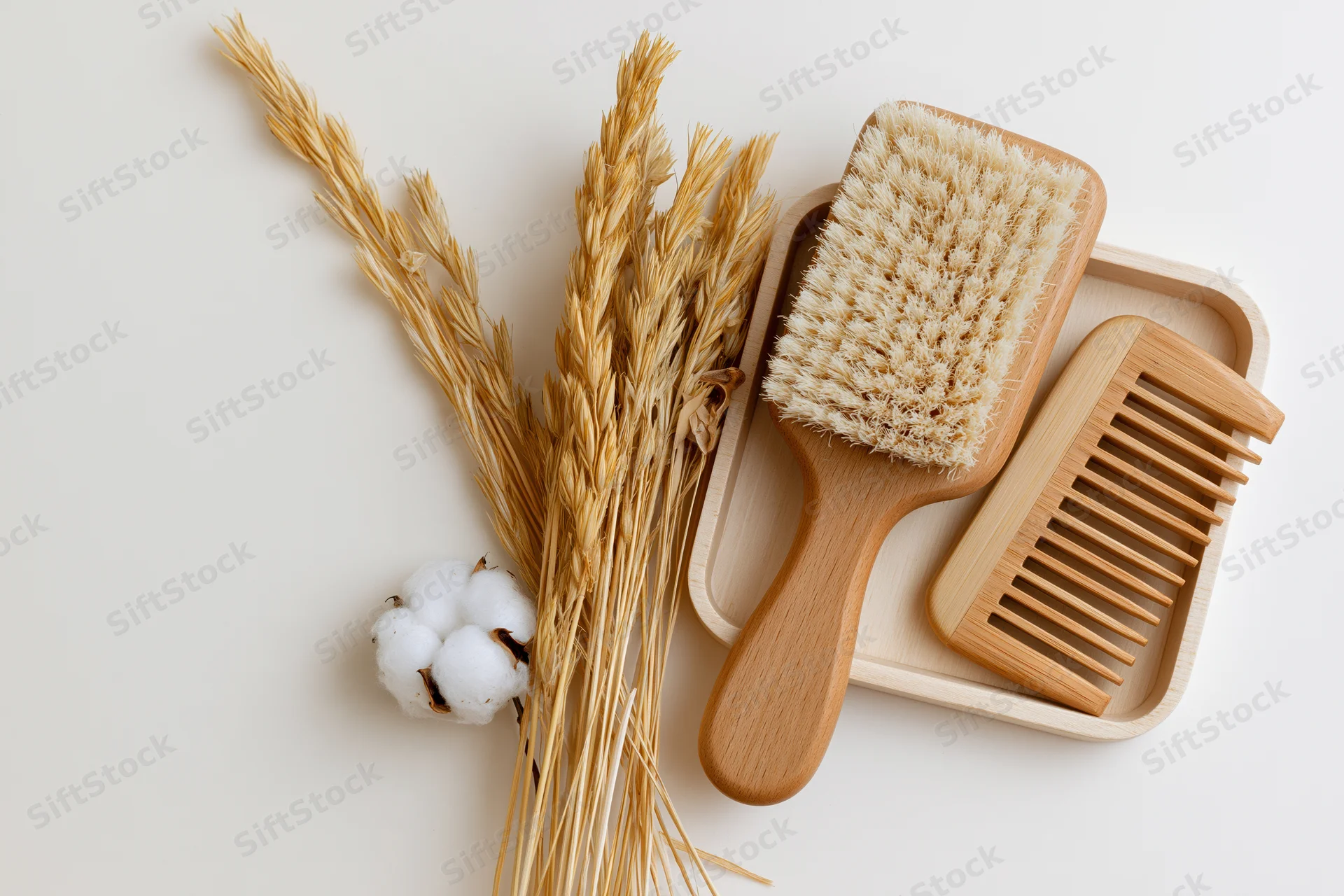 Wooden hairbrush and wide-tooth comb on a tray with dried wheat and cotton, flat lay on white surface