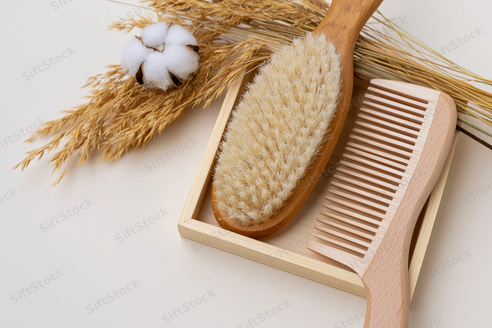 Wooden hairbrush and wide-tooth comb in a bamboo tray with cotton and wheat stalks, flat lay on white surface