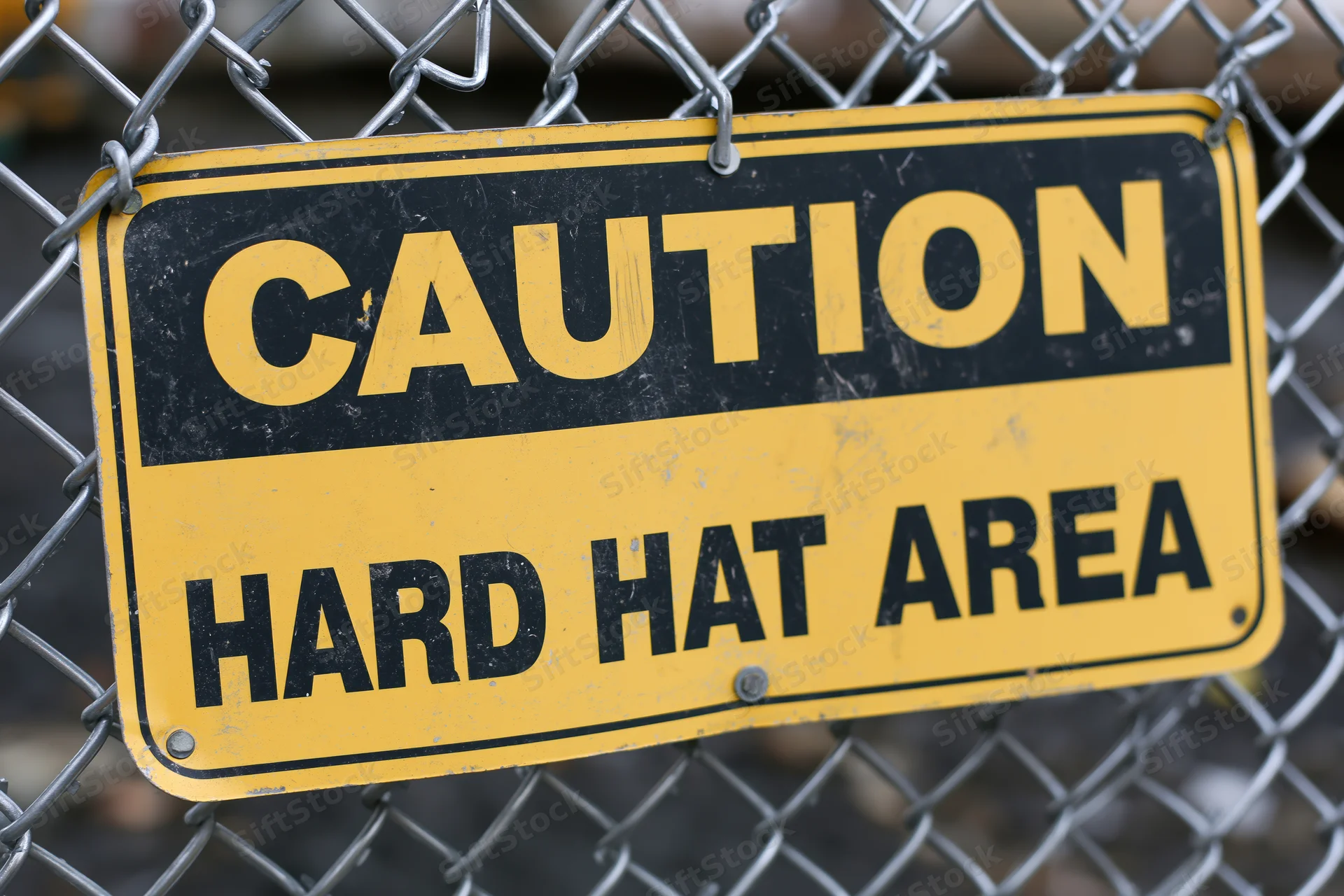 Yellow and black caution hard hat area sign bolted to chainlink fence