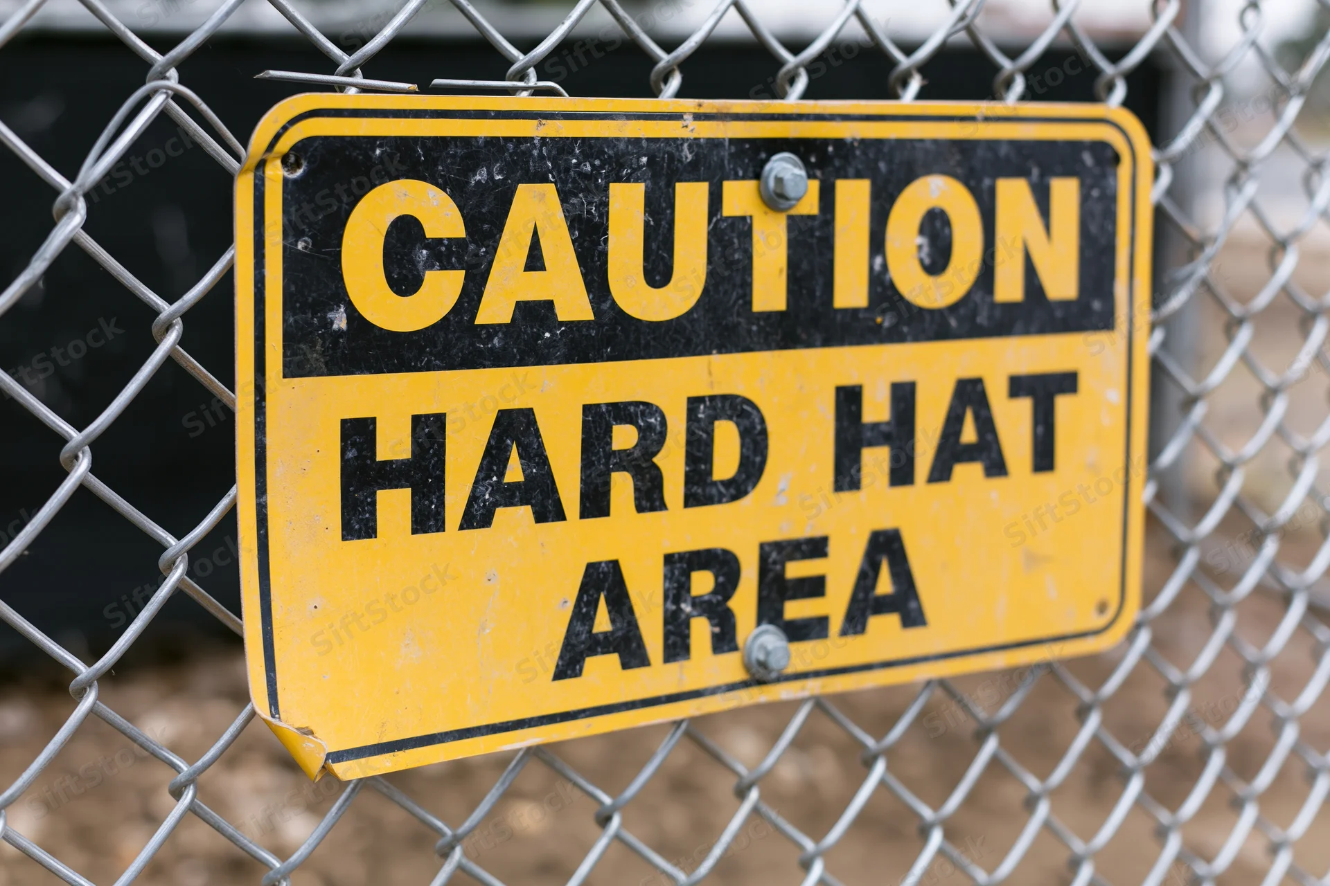 Yellow and black caution hard hat area sign bolted to chainlink fence