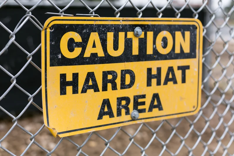 Yellow and black caution hard hat area sign bolted to chainlink fence
