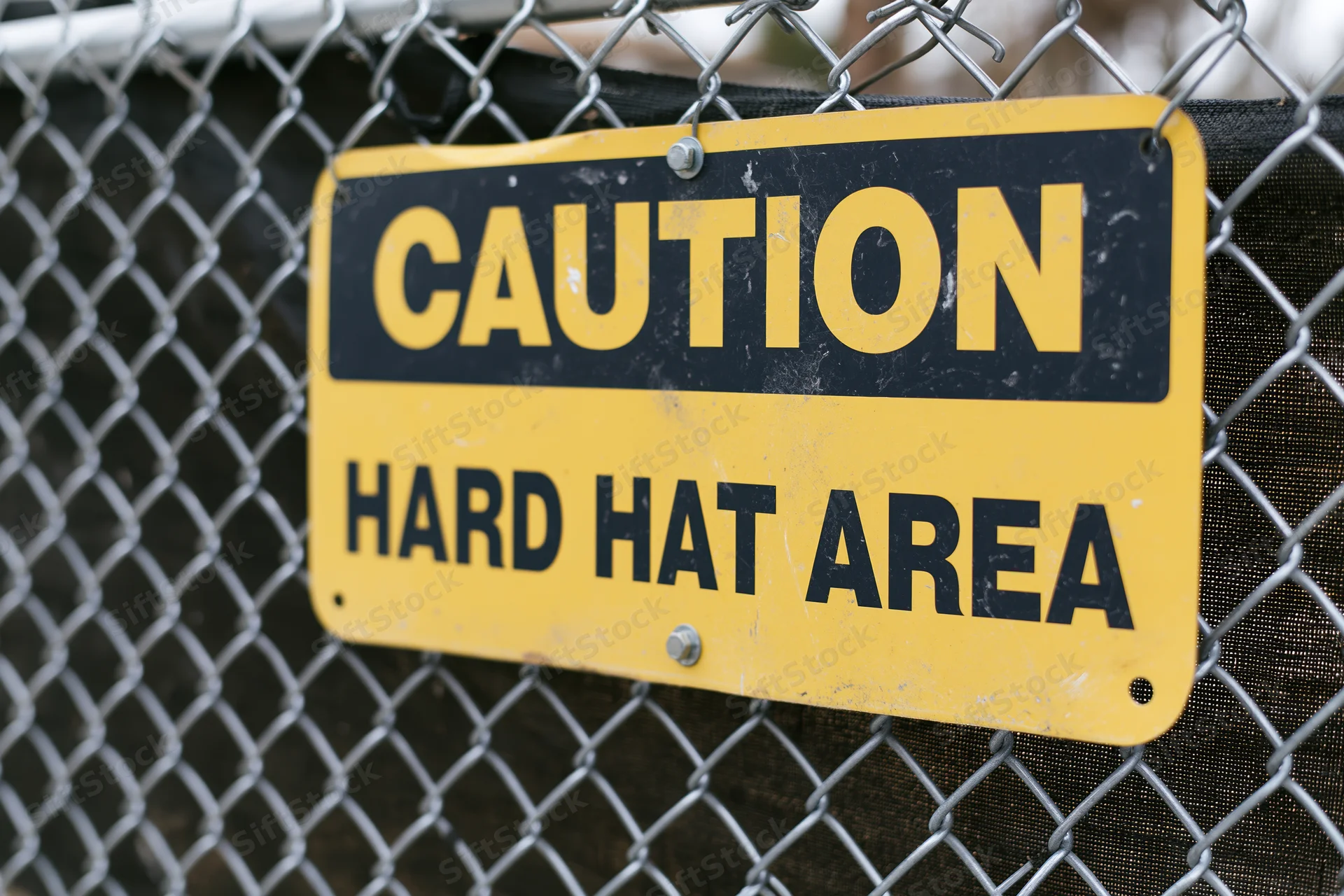 Yellow and black caution hard hat area sign bolted to chainlink fence at construction site