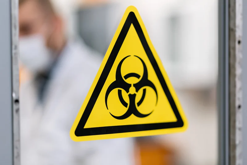 Yellow triangular biohazard warning sign on glass with scientist in lab coat and mask blurred in background