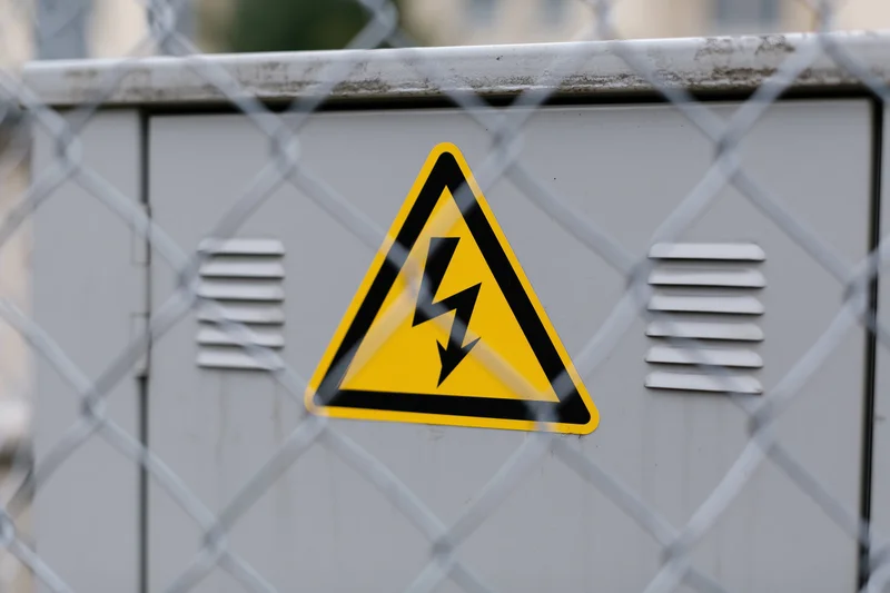 Yellow triangular electrical hazard warning sign on metal cabinet behind chainlink fence