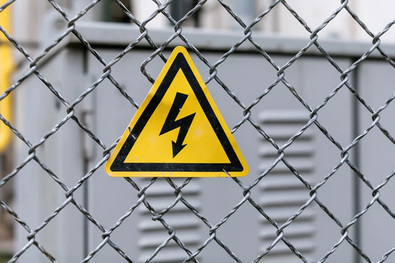 Yellow triangular electrical hazard warning sign attached to chainlink fence in front of industrial equipment
