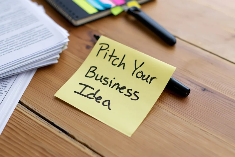 Yellow sticky note with handwritten text pitch your business idea on wooden desk beside documents and planner