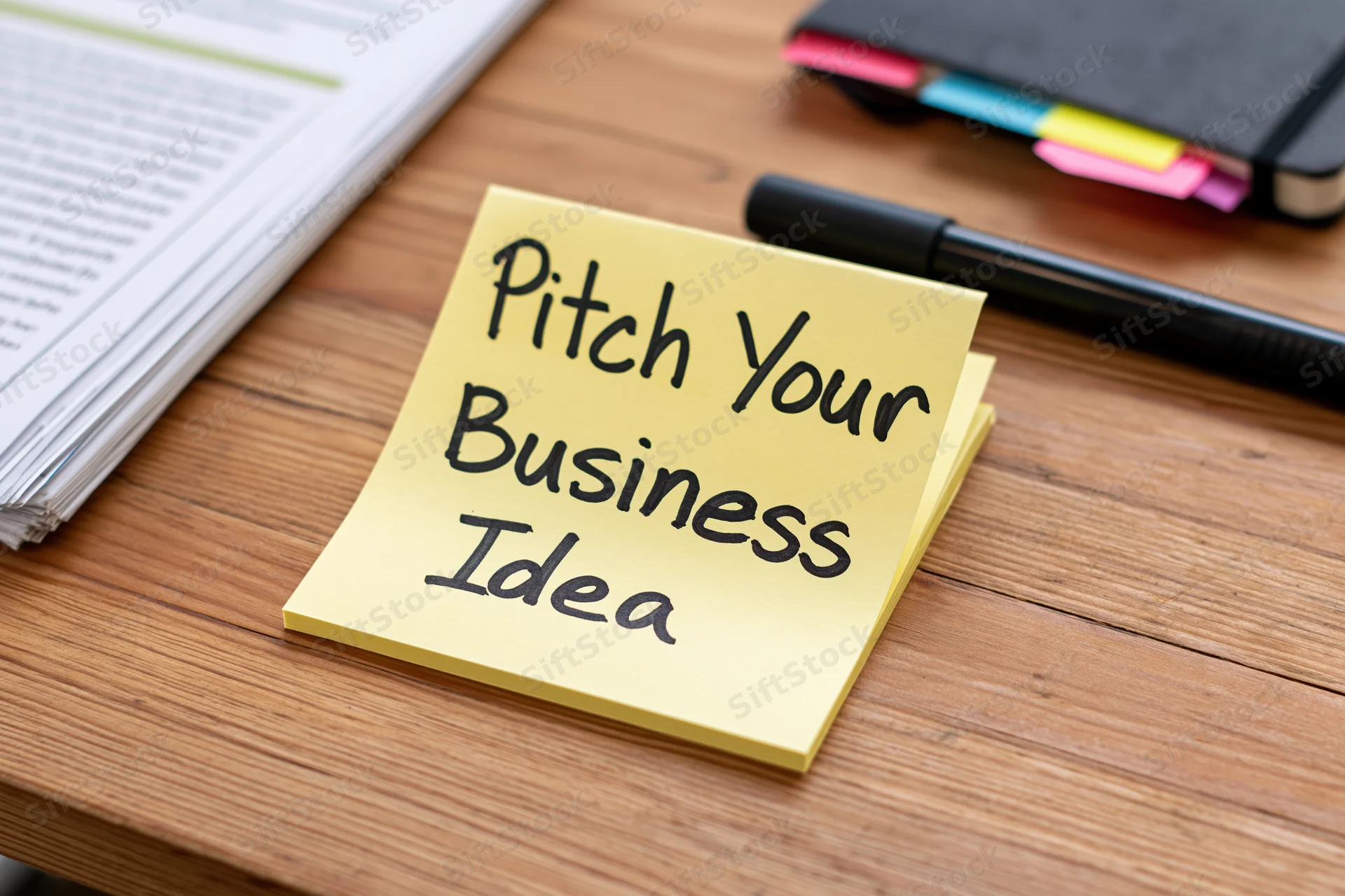 Yellow sticky note with handwritten text reading pitch your business idea on wooden desk