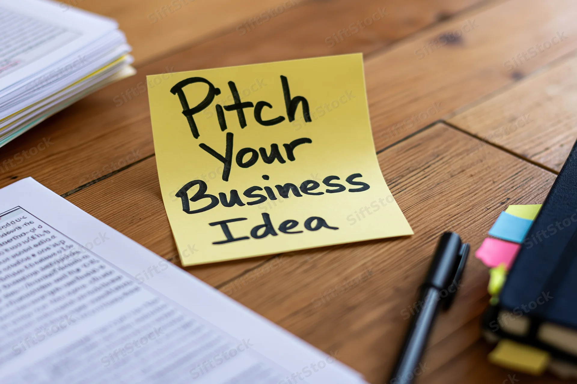 Yellow sticky note with handwritten text reading pitch your business idea on wooden desk surrounded by documents and office supplies