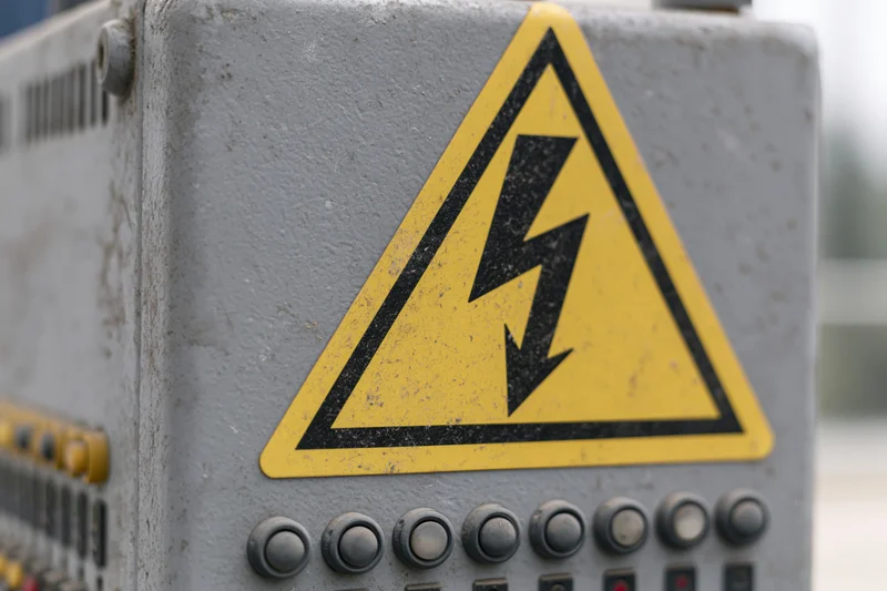 Yellow triangular high-voltage warning sign on a gray industrial control panel with buttons and switches