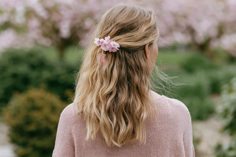 Woman with wavy blonde hair adorned with pink cherry blossoms standing in a blooming garden