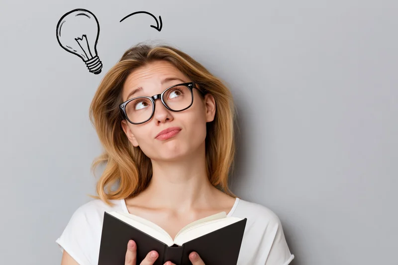 Young woman with black-rimmed glasses holding an open book and looking up at a sketched lightbulb above her head