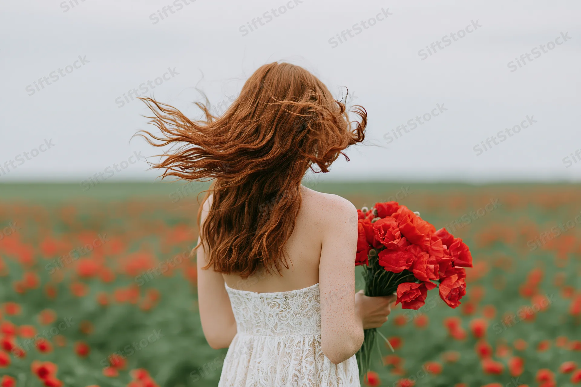 Woman with long red hair blowing in wind holding bouquet of red poppies in field