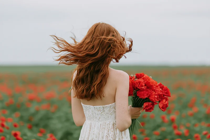 Woman with long red hair blowing in wind holding bouquet of red poppies in field