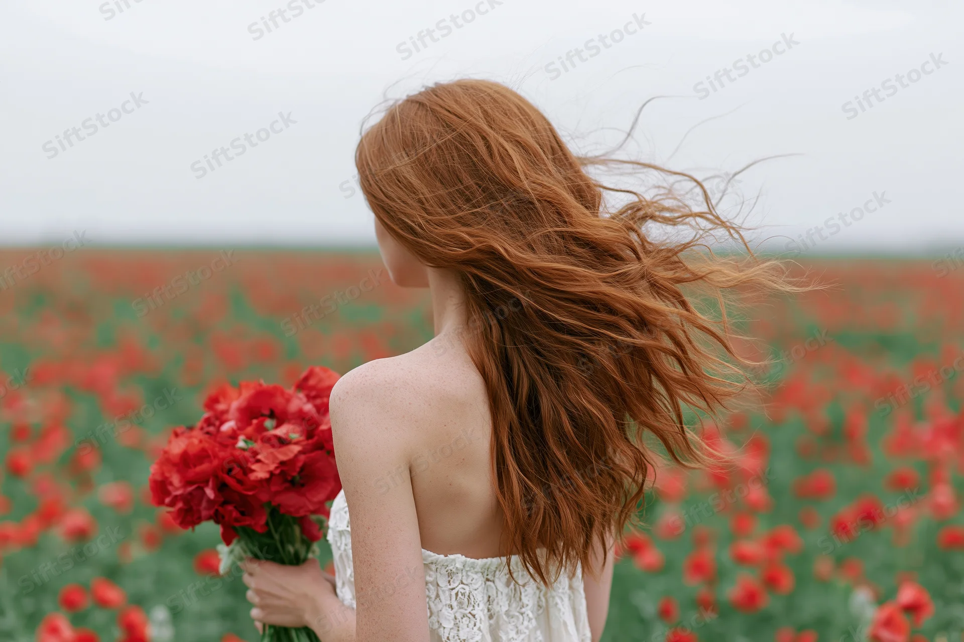 Woman with long auburn hair blowing in wind holding bouquet of red poppies in field