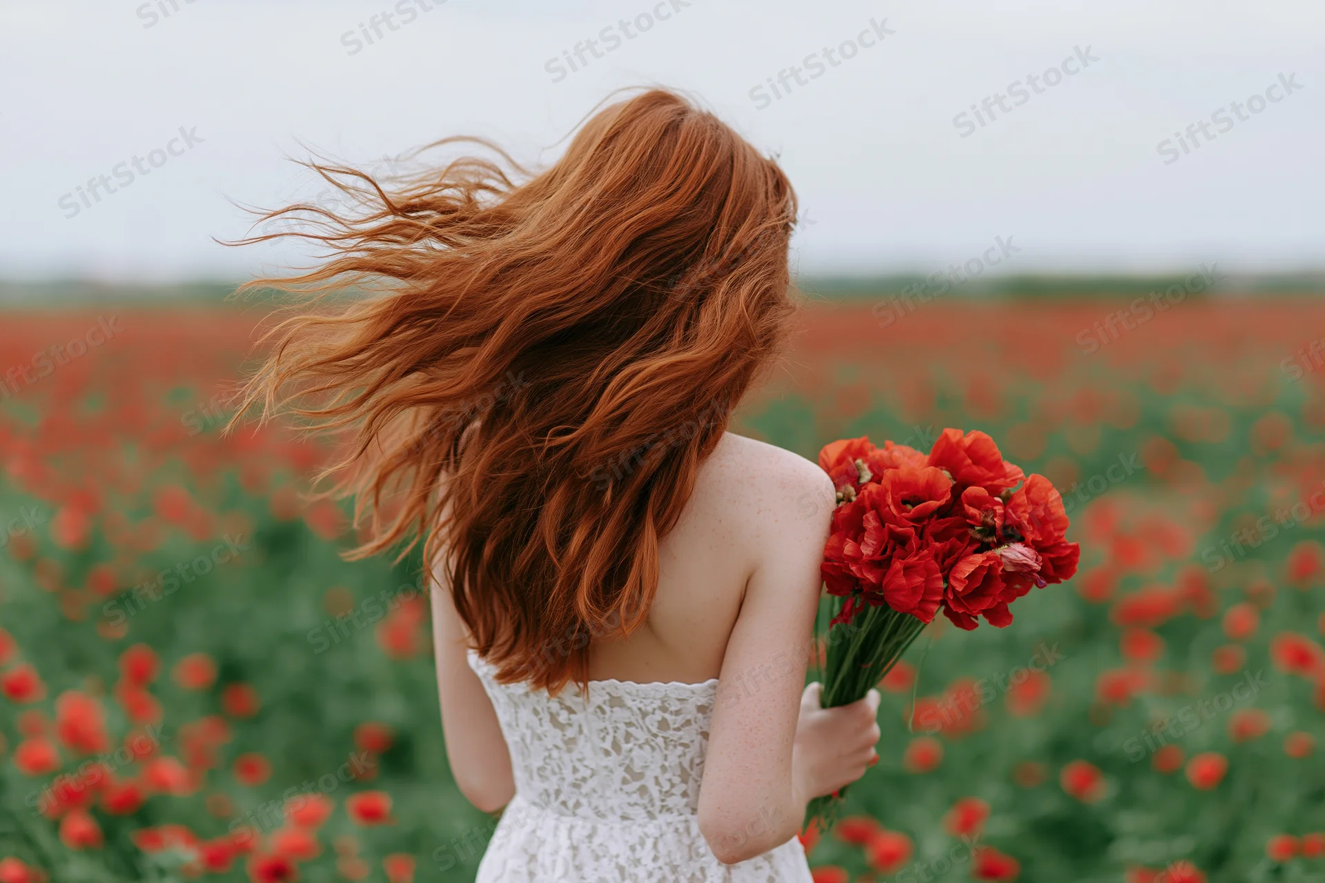 Woman with long red hair blowing in the wind holding a bouquet of red poppies in a field