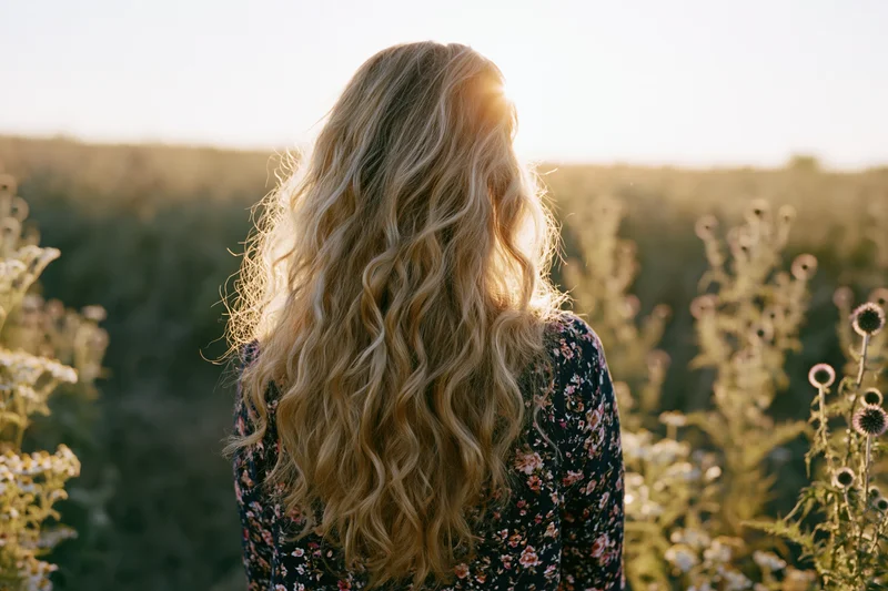 Woman with long wavy blonde hair standing with her back to the camera in a field of wildflowers at sunset