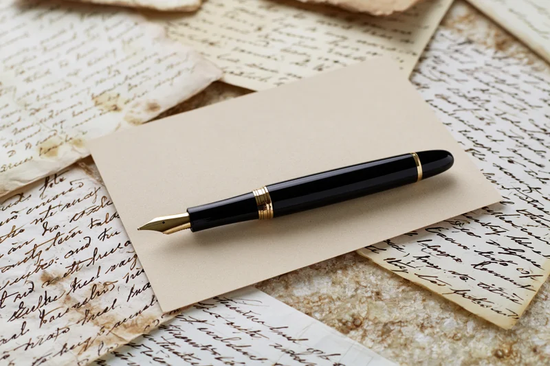 Black and gold fountain pen resting diagonally on a blank envelope atop aged handwritten letters