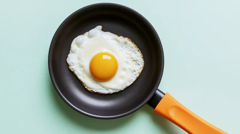 Fried sunny-side-up egg in a small dark nonstick frying pan on a mint green surface