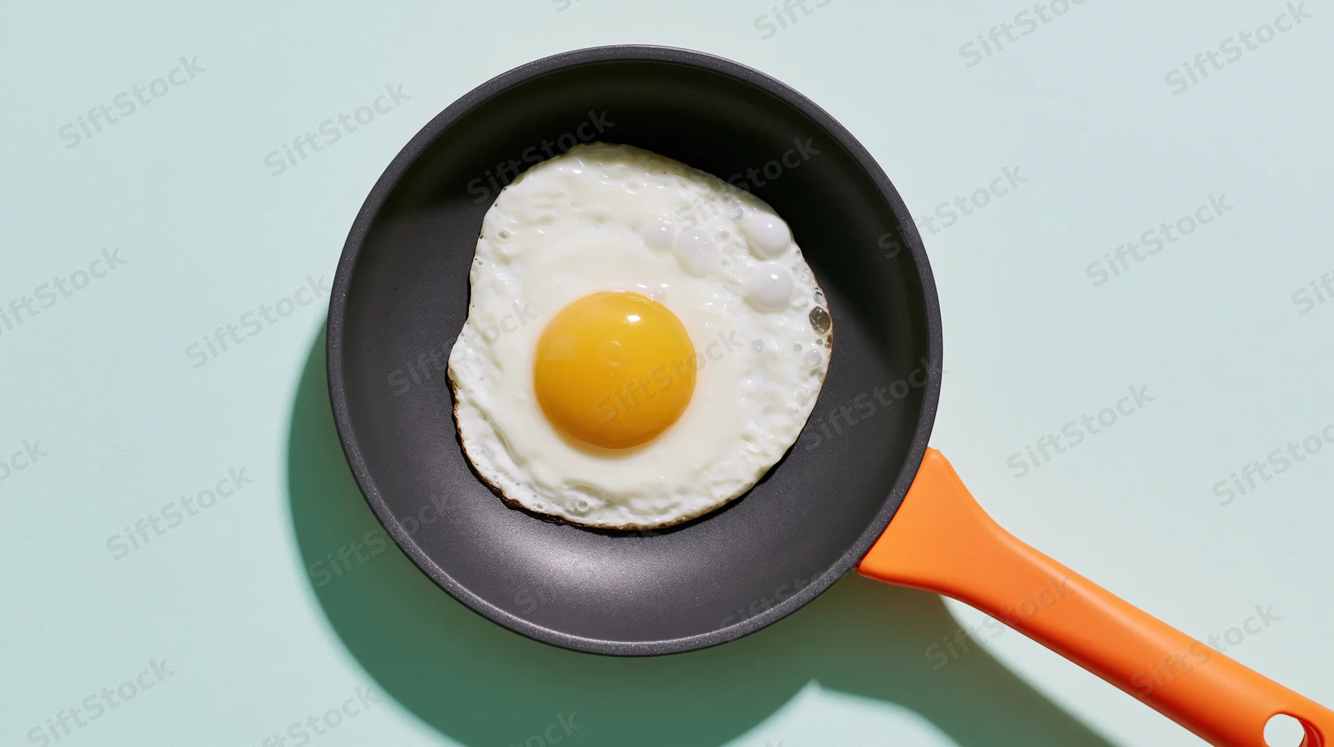 Fried sunny-side-up egg in a small dark nonstick pan with orange handle on mint background