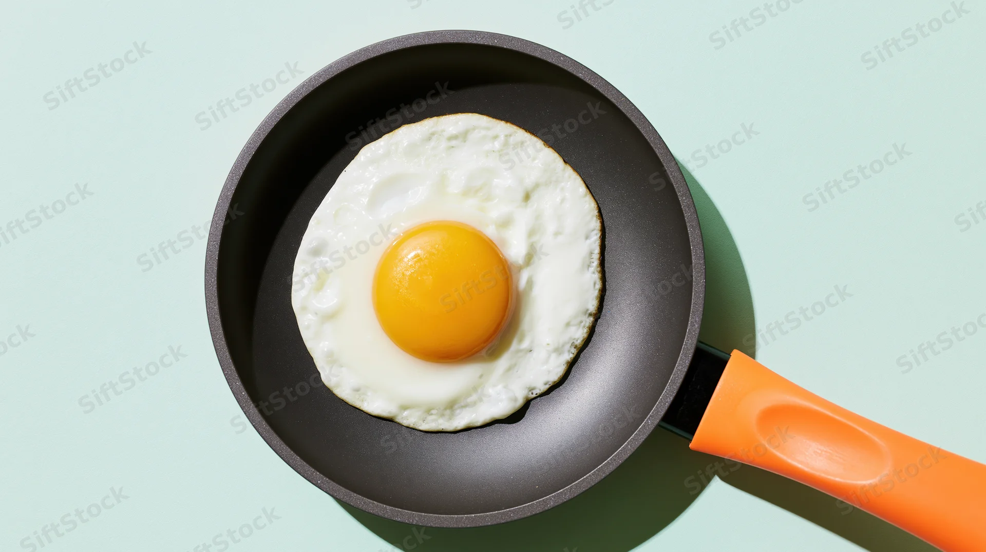 Fried sunny-side-up egg in a small dark nonstick frying pan with orange handle on light background