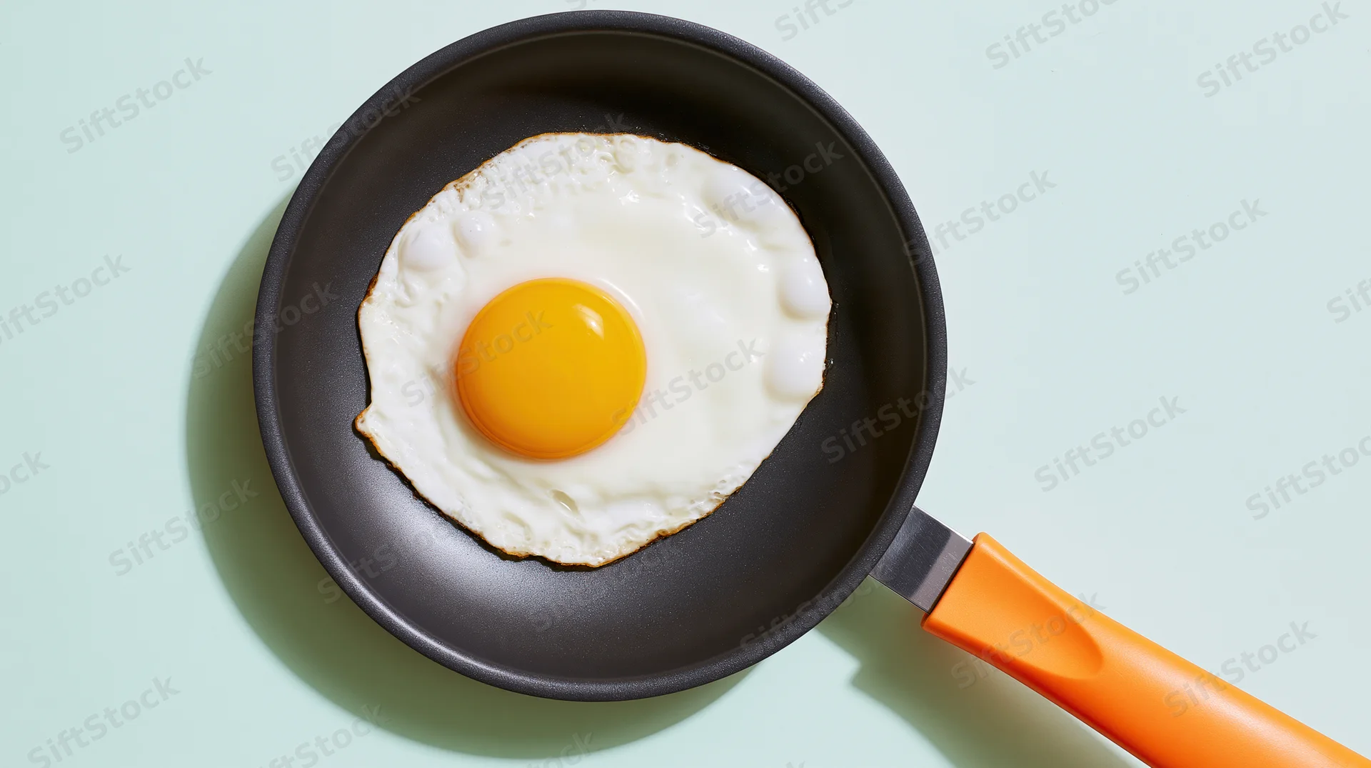 Fried sunny-side-up egg in a small dark nonstick frying pan with orange handle on light green surface