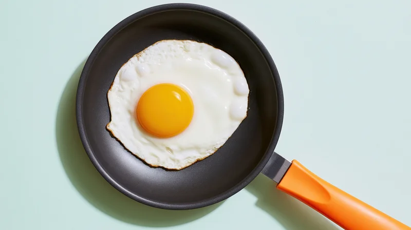 Fried sunny-side-up egg in a small dark nonstick frying pan with orange handle on light green surface