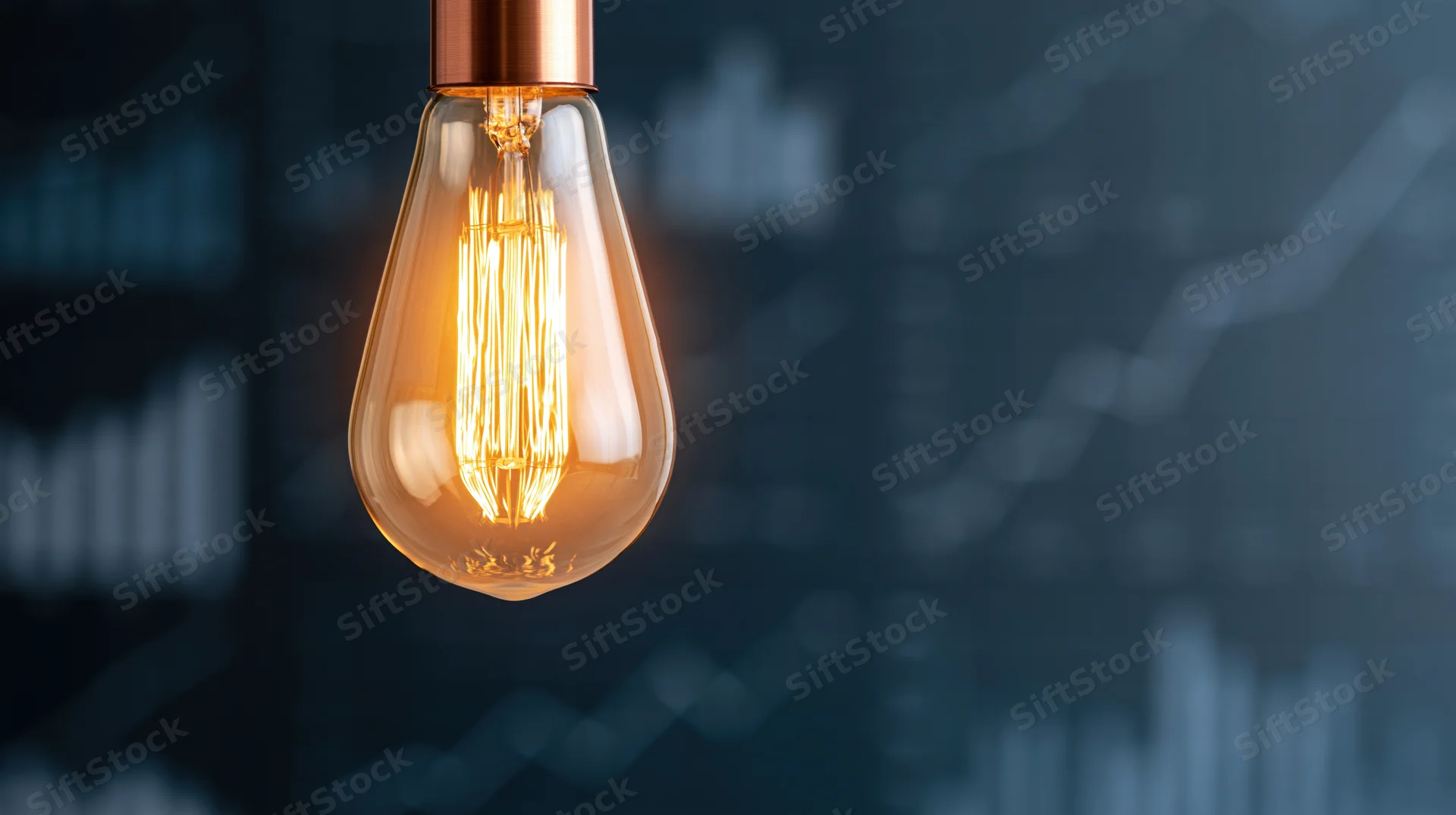 Glowing incandescent lightbulb with visible filament against a blurred background of financial charts and graphs