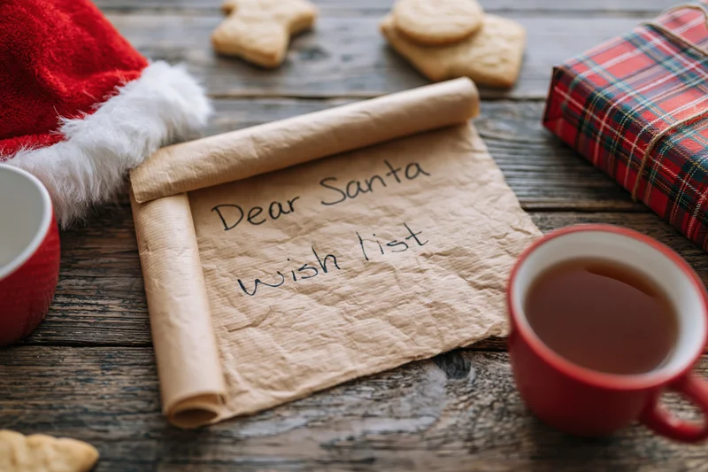 Handwritten wish list on aged kraft paper scroll with santa hat, cookies and tea on wooden table