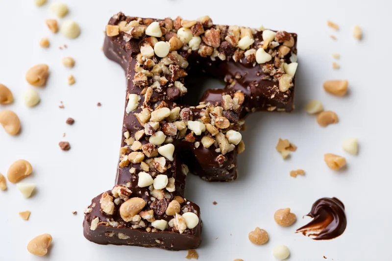 Chocolate letter f shaped candy covered in crushed nuts and white chocolate pieces on white surface