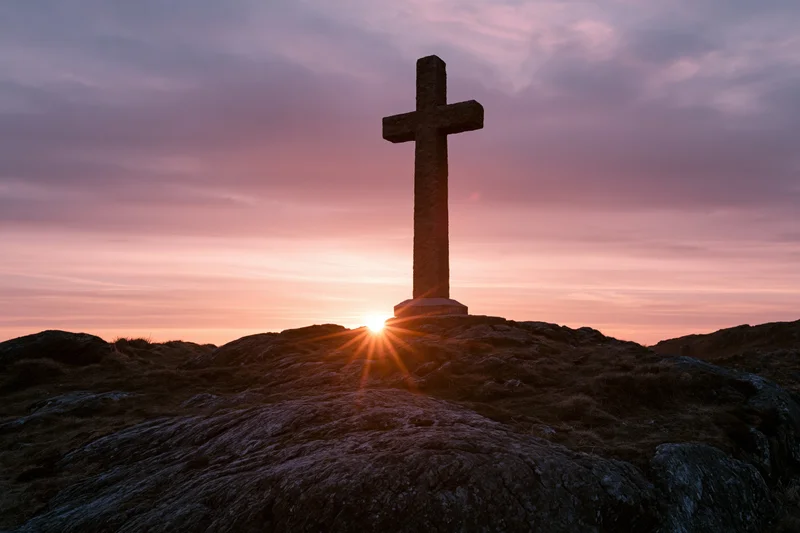 Stone cross silhouetted on rocky hilltop at sunset
