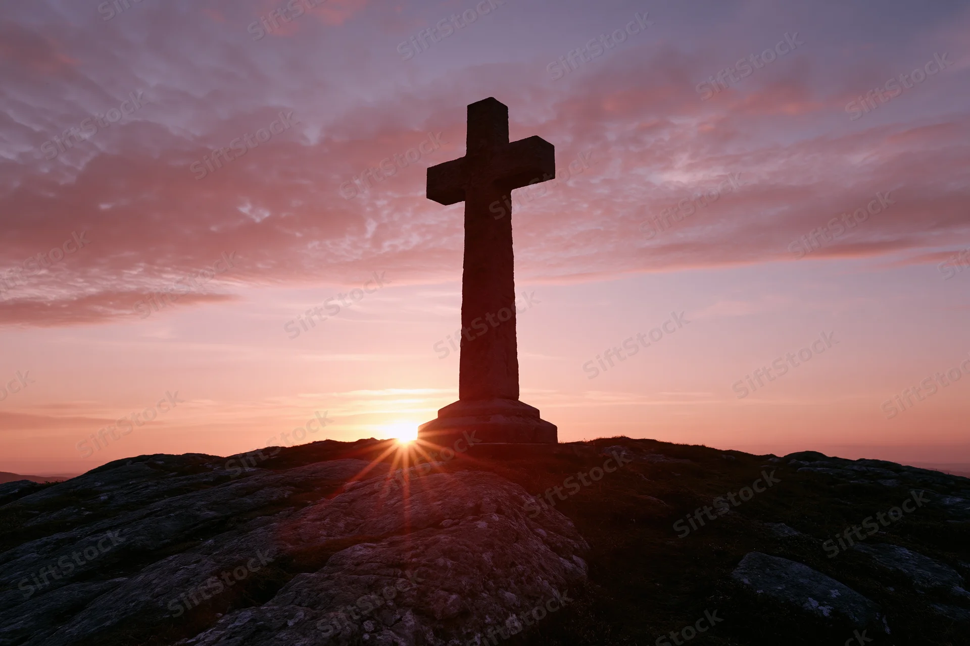 Stone cross silhouetted on rocky hilltop at sunset