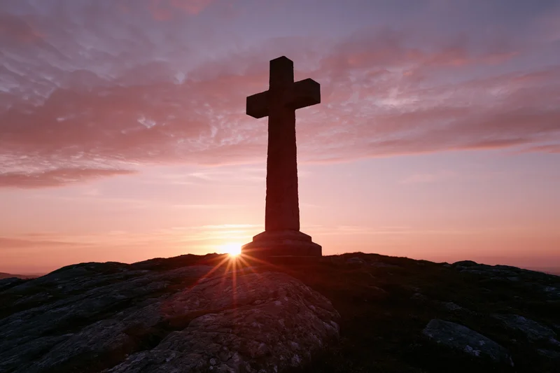 Stone cross silhouetted on rocky hilltop at sunset
