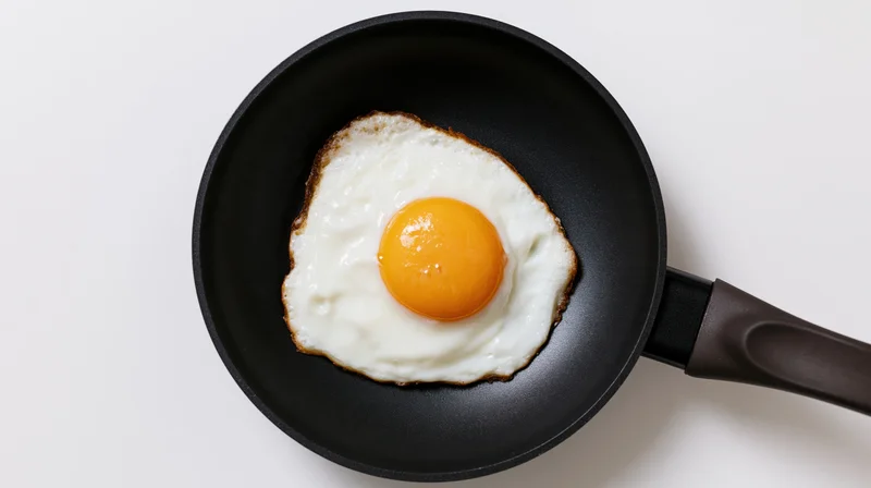 Single fried egg with set yolk in a small black nonstick pan on white surface