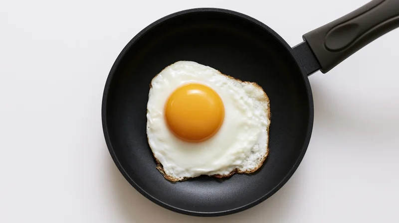 Single fried egg with set yolk in small black nonstick skillet on white surface
