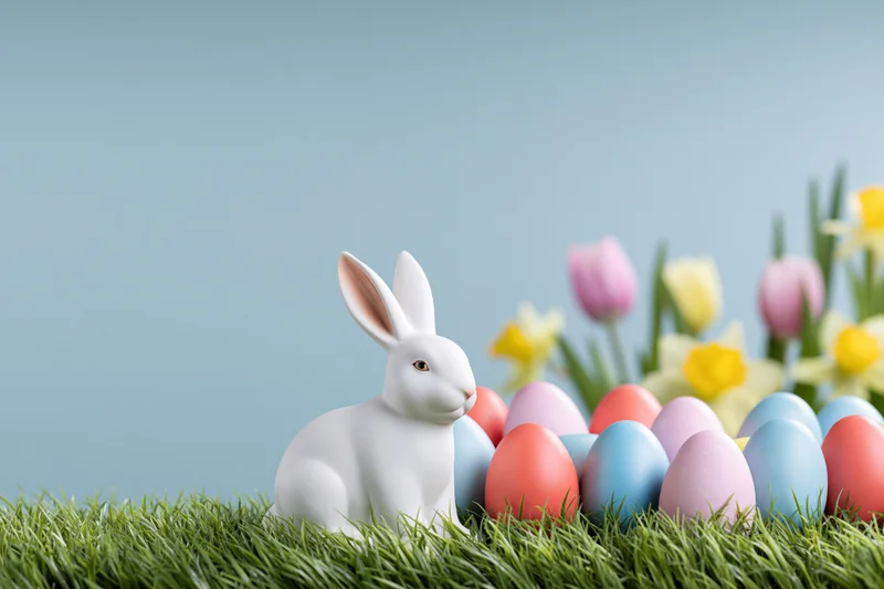 White ceramic easter bunny figurine sitting among pastel eggs and spring flowers on green grass