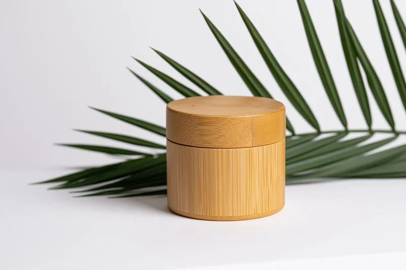 Cylindrical bamboo cosmetic jar with fitted lid on white surface beside palm leaf
