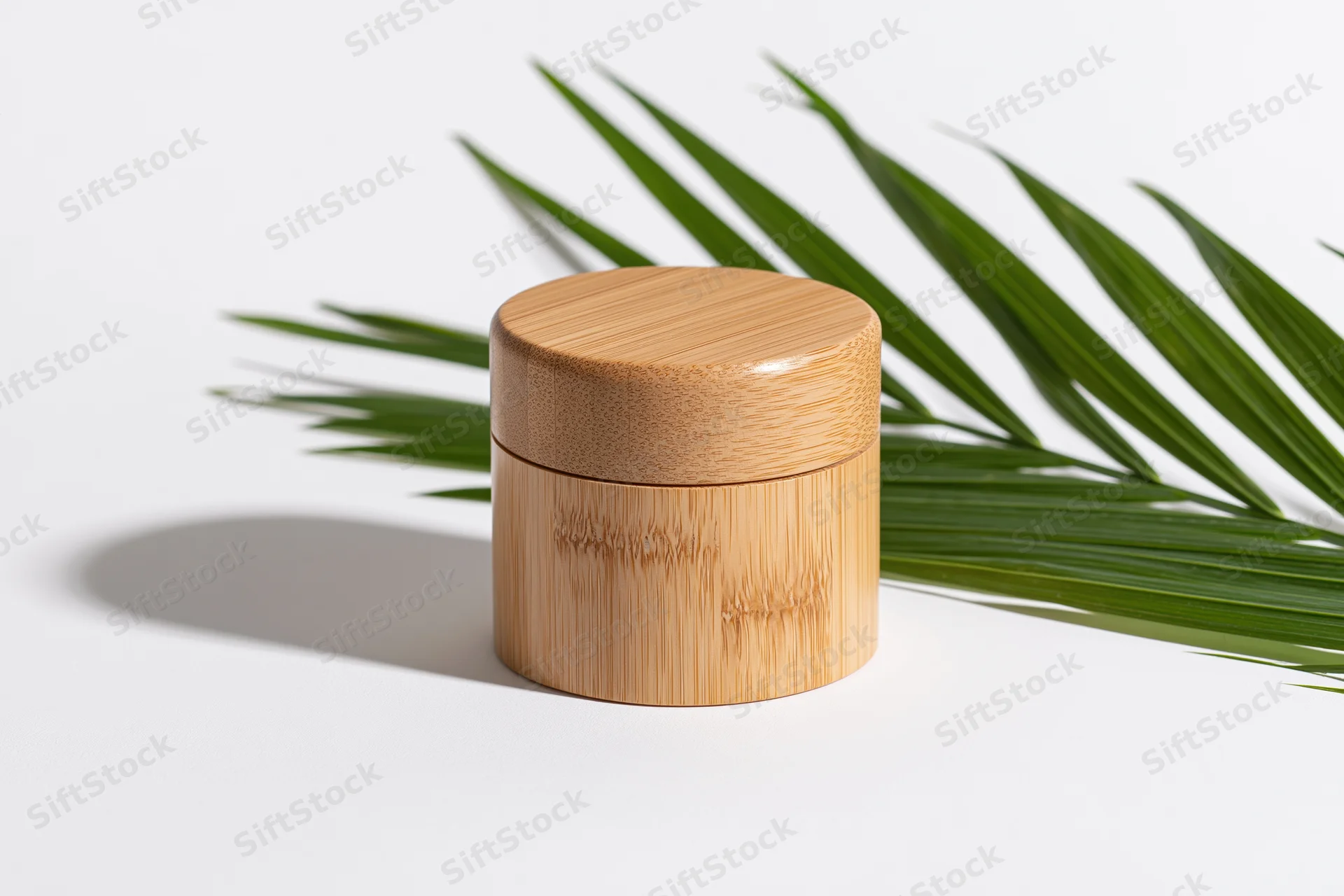 Small cylindrical bamboo cosmetic jar with fitted lid on white surface beside green palm leaf