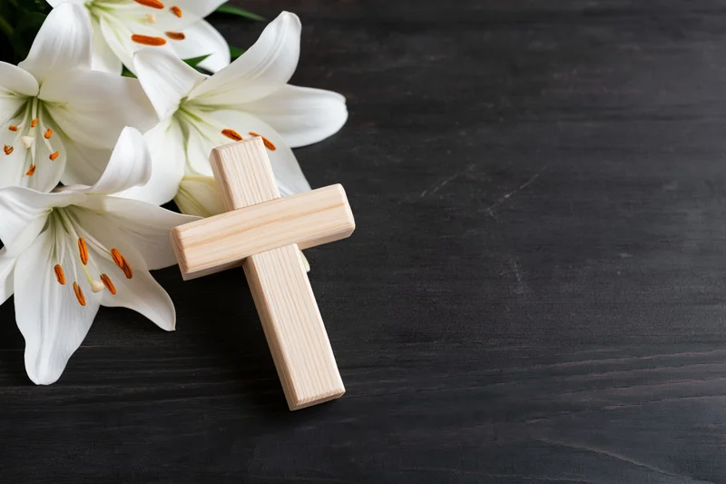 Wooden cross leaning against white lilies on dark wooden surface