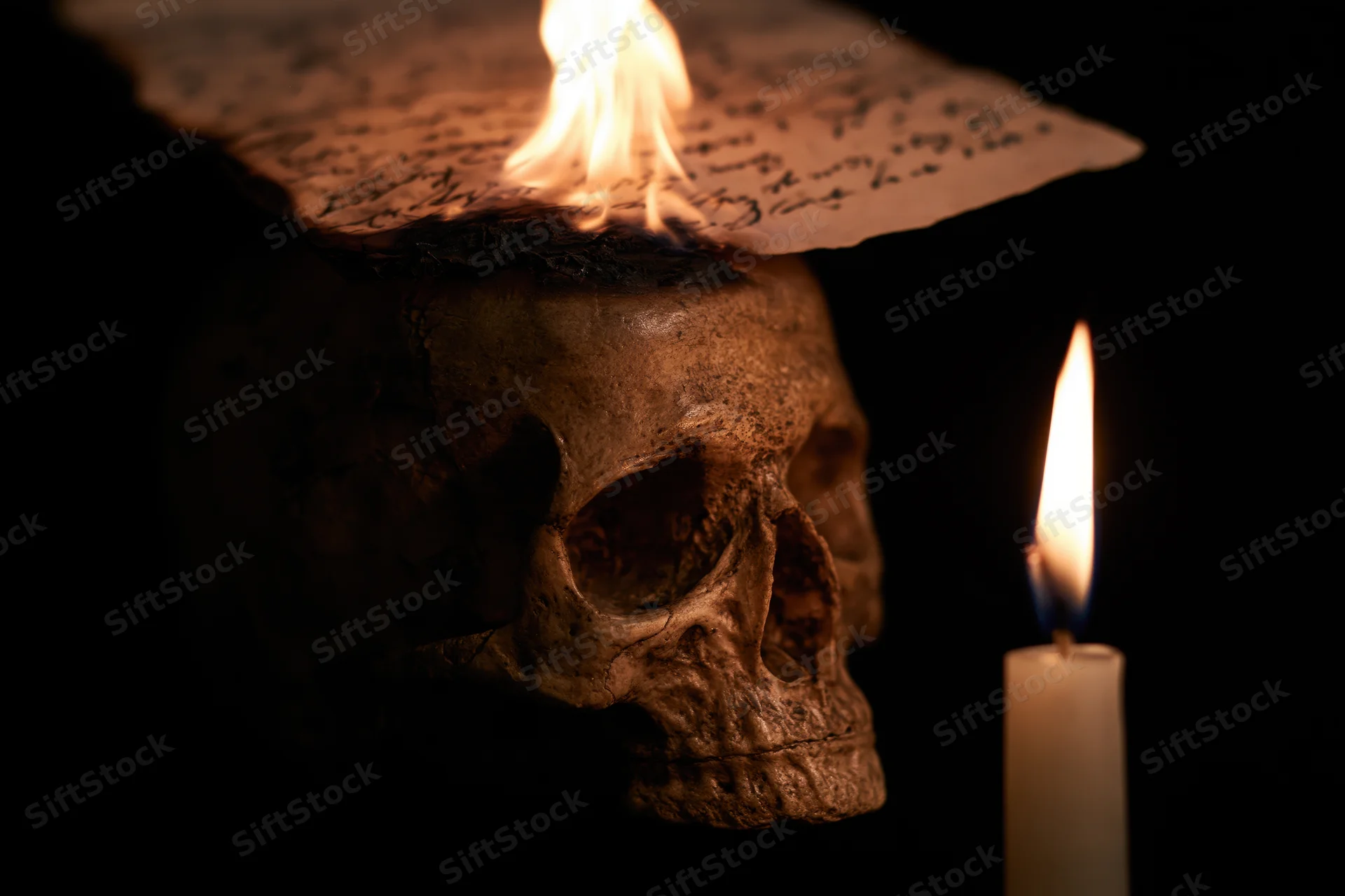 Weathered human skull with burning paper on top and lit candle beside it in dark setting