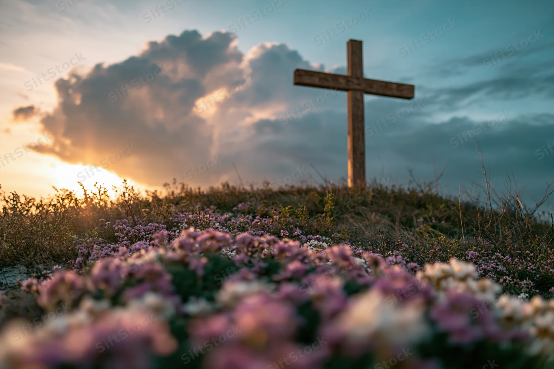 Wooden cross on a grassy hilltop at sunset surrounded by wildflowers