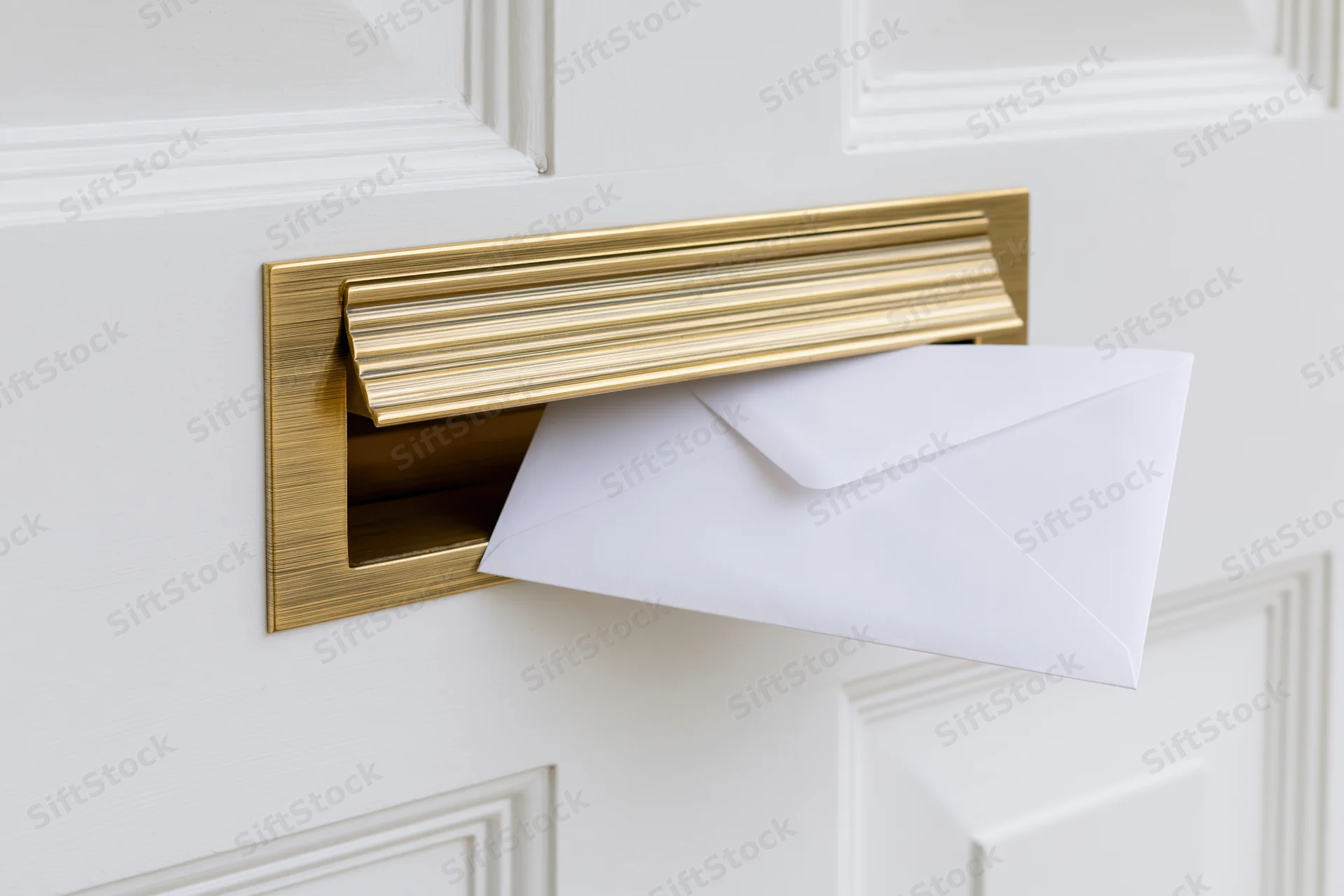 White envelope partially inserted into brass mail slot on white door