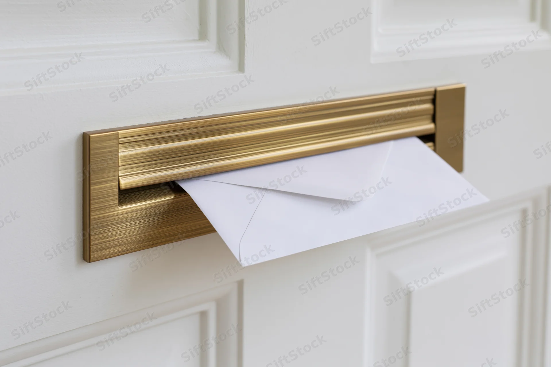 White envelope partially inserted into brass mail slot on white door
