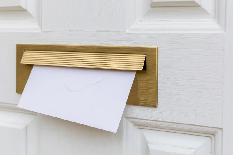 White envelope partially inserted into brass mailbox on white door