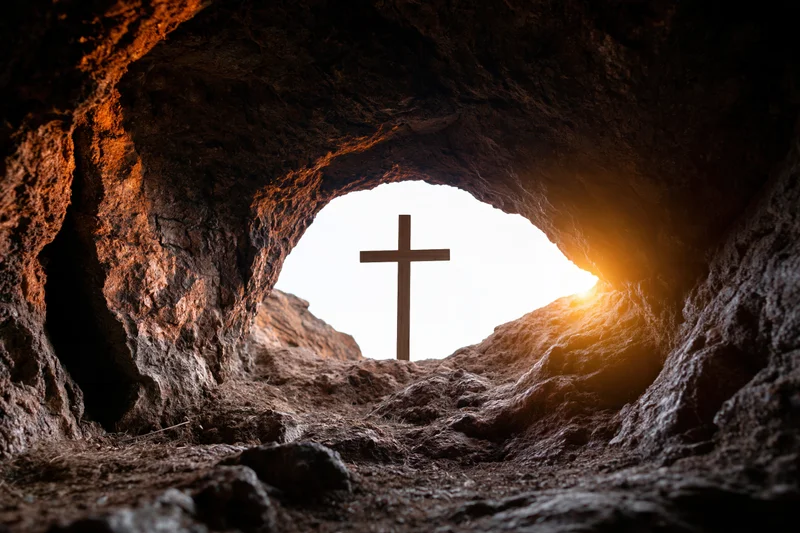 Wooden cross silhouetted at cave entrance with sunlight streaming through rocky opening