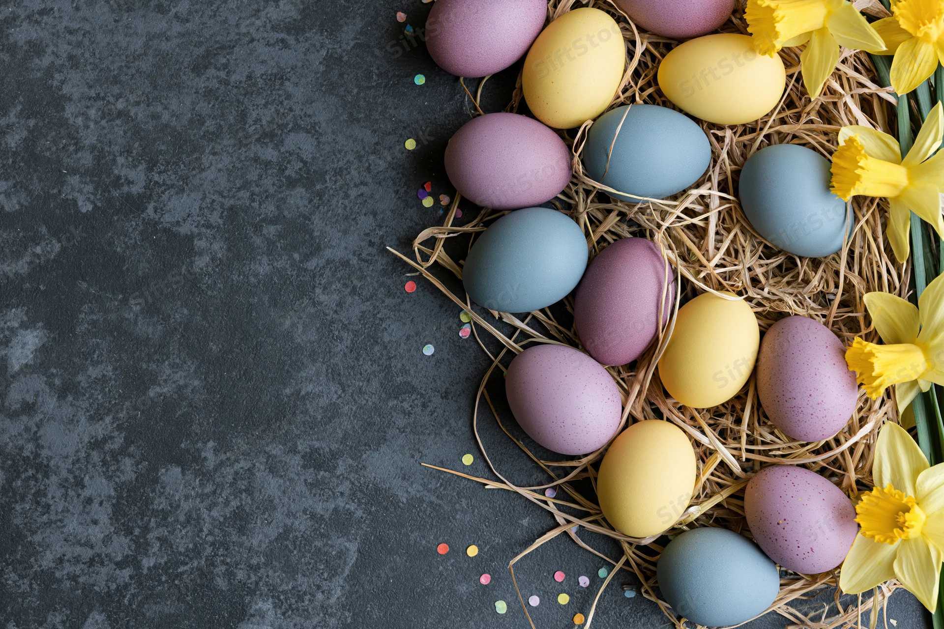 Pastel and jewel-toned easter eggs arranged in a straw nest with yellow daffodils on a dark textured surface