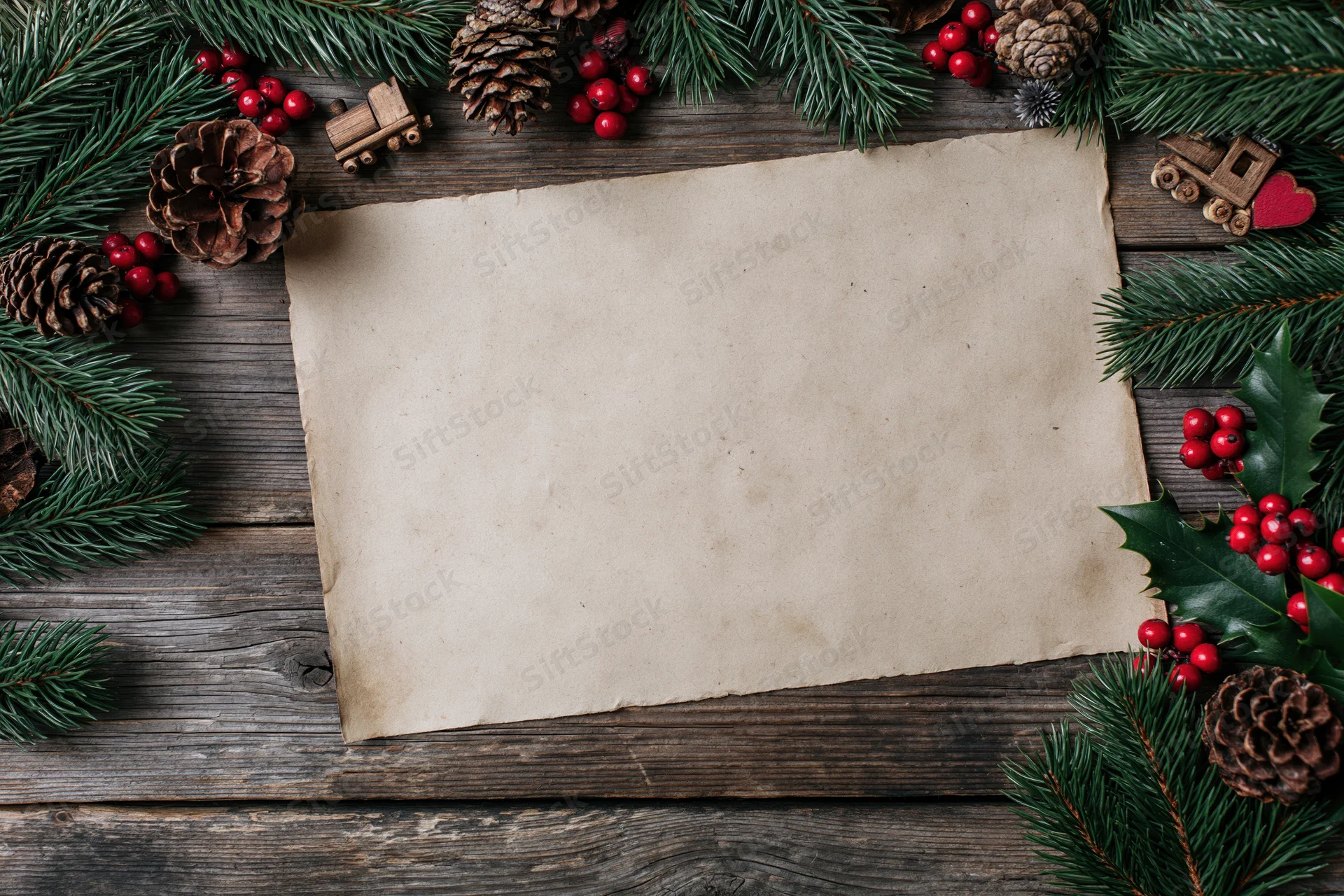 Blank aged paper sheet on wooden surface framed by pine branches, holly, berries and pinecones for christmas decoration